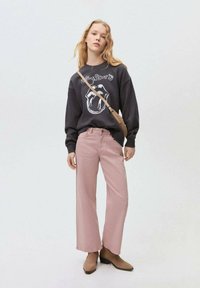 Svart sweatshirt med logotyp, rosa vida byxor och bruna klackade stövletter. Modellen bär en axelremsväska. Neutral bakgrund.