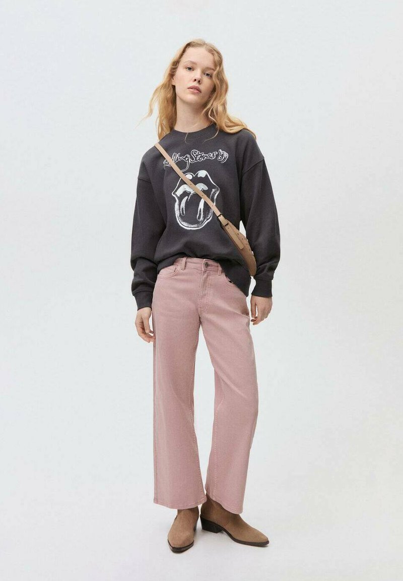 Svart sweatshirt med logotyp, rosa vida byxor och bruna klackade stövletter. Modellen bär en axelremsväska. Neutral bakgrund.