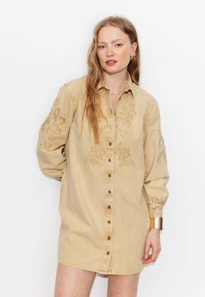 Jeune femme aux longs cheveux portant une robe beige boutonnée avec broderie florale et un bracelet manchette doré, debout devant un fond blanc.