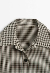 Chemise à col avec motif à petits carreaux noir, blanc et marron, un seul bouton noir fermé au centre devant.