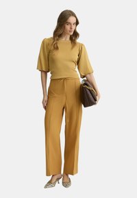 Top testurizzato giallo senape con maniche a sbuffo, abbinato a pantaloni a gamba larga e vita alta. Una borsa marrone e scarpe a fantasia completano il look.