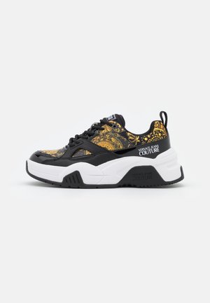 Schwarzer und goldener Sneaker mit dicker weißer Sohle, "Versace Jeans Couture"-Branding an der Seite und Fersenlasche, von rechts gezeigt.