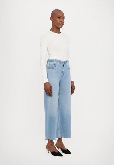 Šviesiai mėlyni laisvo kirpimo džinsai su neapdirbtu kraštu, derinami su prigludusia balta ilgomis rankovėmis megztine ir juodais aukštakulniais be užsegimo.