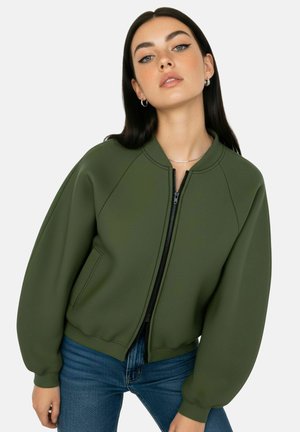 Jeune femme aux longs cheveux foncés portant des boucles d'oreilles argentées en forme de cercle, une veste bomber zippée vert olive et un jean bleu, posant sur fond blanc.