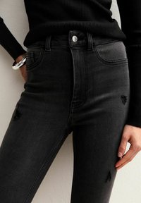 Vaqueros de mezclilla negros de tiro alto con un ajuste slim, que presentan detalles desgastados y un diseño clásico de cinco bolsillos. Textura suave con un ligero brillo.