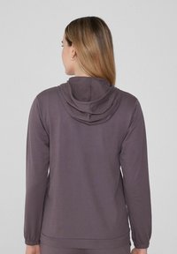 Sudadera con capucha de manga larga en un tejido de color púrpura medio. Presenta puños elásticos y un corte amplio. Textura suave con detalles en la capucha de capas.