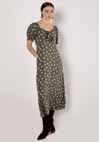 Robe midi vert olive avec des manches courtes bouffantes et un décolleté carré. Imprimé floral blanc sur l'ensemble, avec un détail à nouer et des poches latérales.