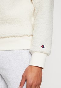 Krämsfleece-tröja med texturerad tyg, ribbade ärmslut och broderad logotyp på ärmen. Matcher med ljusgråa sweatpants.