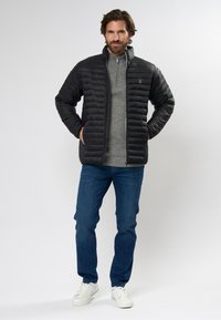 Schwarze Pufferjacke mit gestepptem Design, grauer Reißverschluss-Pullover darunter, blaue Jeans und weiße Sneakers. Mit Fronttaschen.