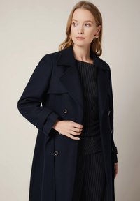 Navy blue trench coat z grubego materiału, zapinany na podwójne guziki metalowe, z szerokim kołnierzem i zwiniętymi mankietami, połączony z górą w paski.