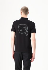 Polo nero a maniche corte, con un design di linee bianche che rappresenta un volto con occhiali da sole sulla schiena. Materiale in cotone morbido.