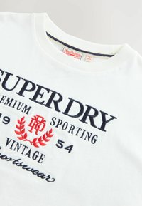 Bílý bavlněný tričko s námořnickým modrým a červeným vyšitým textem. Kola krk, krátké rukávy a vintage styl grafiky. Měkká textura.