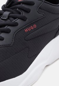 Gros plan sur une basket noire en tissu texturé, lacets noirs, logo rouge "HUGO" et semelle blanche épaisse.