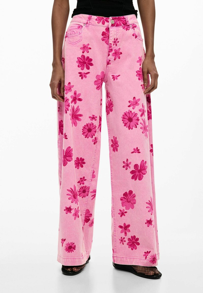 Persona vistiendo jeans rosa de tiro alto y pierna ancha con estampado floral en tonos rosa oscuro y zapatos negros de red sobre fondo blanco.