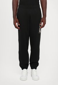 Pantalones de chándal negros con cintura y puños elásticos, que presentan un acento de logo en el lateral. El material tiene una textura suave, combinados con zapatillas blancas.