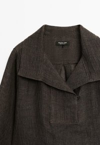 Chemise en tissu texturé brun avec un large col, une fermeture à boutons et une poche avant, conçue pour une coupe ample. Étiquette visible à l'intérieur.