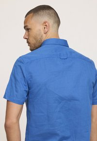 Chemise bleue à manches courtes avec boutons, fabriquée en tissu léger, dotée d'un col classique et d'un détail au dos. Texture douce.