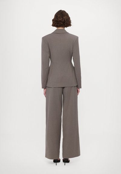 Braun maßgeschneiderte Blazerjacke mit Schulterpolstern und passender weit geschnittener Hose, aus glattem Stoff gefertigt. Styliert mit High-Heels.