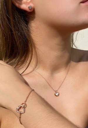 L'ensemble de bijoux en or rose comprend des boucles d'oreilles en forme de cœur, un collier avec un pendentif en forme de cœur présentant une pierre centrale, et un bracelet avec des accents en forme de cœur.