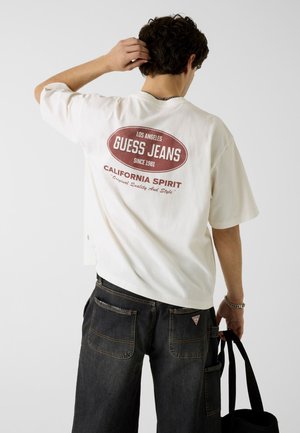 Jeune homme portant un T-shirt Guess Jeans blanc avec logo rouge et jean foncé, tenant un sac noir et se grattant l’arrière de la tête.