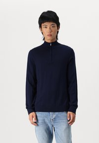 Suéter de punto azul marino con cremallera, cuello alto y mangas largas, con una textura suave y puños acanalados. Se lleva con jeans de mezclilla claros.