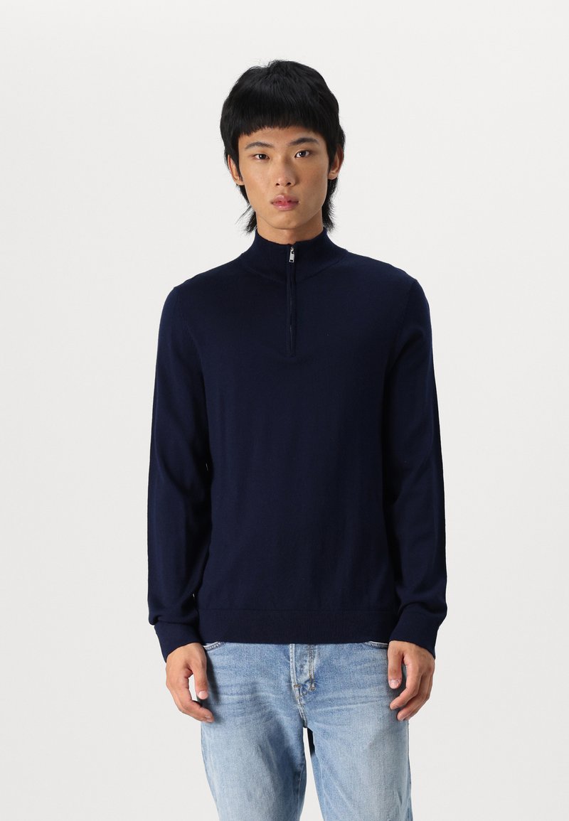 Suéter de punto azul marino con cremallera, cuello alto y mangas largas, con una textura suave y puños acanalados. Se lleva con jeans de mezclilla claros.