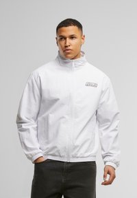 White zip-up jacket with a high collar, elastic cuffs, and a logo patch on the left chest. Lightweight fabric with a smooth texture.