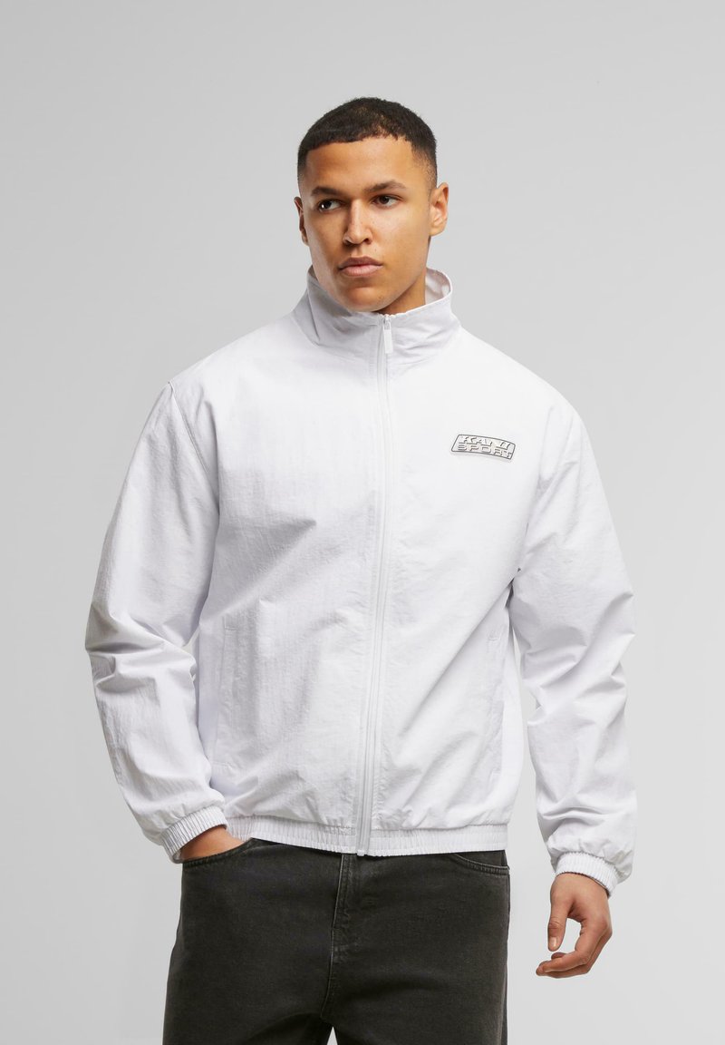 White zip-up jacket with a high collar, elastic cuffs, and a logo patch on the left chest. Lightweight fabric with a smooth texture.