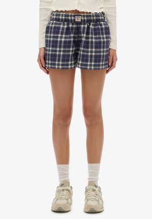 Mujer vistiendo pantalones cortos de cuadros azules y blancos, camiseta de manga larga blanca acanalada, calcetines blancos y zapatillas beige, de pie con las manos relajadas a los lados.