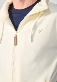 Hvid fleece zip-up hoodie med et navy indre lag, der har et guldbroderet logo, struktureret stof og en læder zippertrækkende detalje.