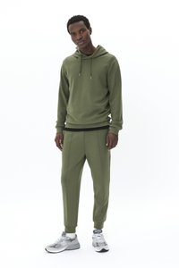 Oliwkowa bluza z kapturem o luźnym kroju i sznurkiem, zestawiona z dopasowanymi zwężanymi spodniami z pionowymi przeszyciami. Noszona z szarymi sneakersami.