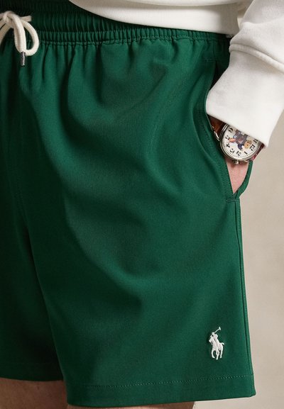 Short vert à cordon avec logo cheval brodé blanc, porté avec une manche blanche, main dans la poche, montrant une montre avec un cadran graphique.