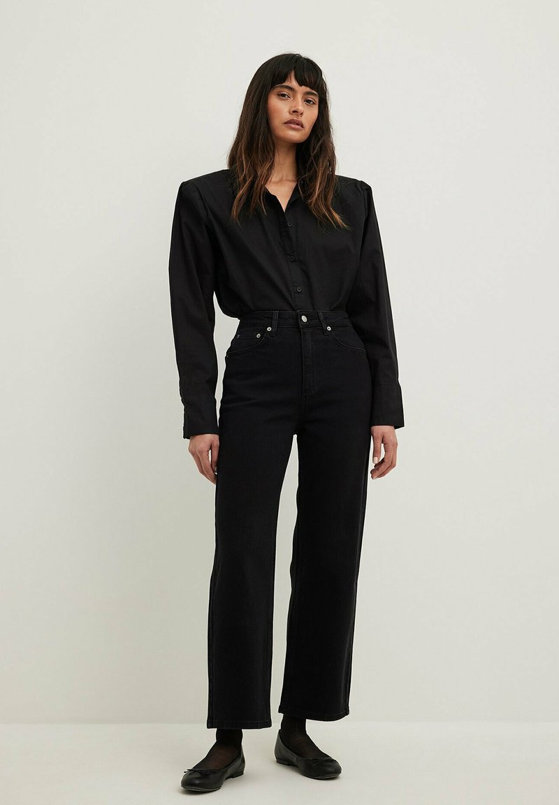 Svart skjorta med knappar och lång ärm, kombinerad med högmidjade svarta jeans och enkla svarta lägenheter, på en neutral bakgrund.
