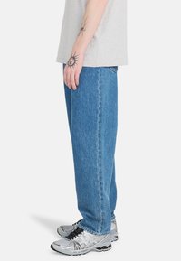 Jeans de denim azul con un corte holgado, piernas rectas y un desvanecimiento sutil. Se llevan con zapatillas deportivas plateadas. Camiseta gris simple visible.
