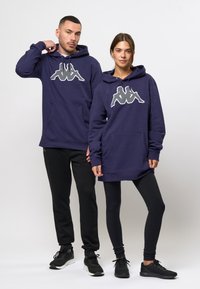 Sudaderas marinas con un gran logo gris, que cuentan con bolsillos frontales. El modelo masculino lleva joggers negros; la modelo femenina tiene leggings.