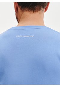 Jasnoniebieska bawełniana koszulka z okrągłym dekoltem i białym logo "PAKO LORENTE" na plecach w pobliżu kołnierza. Gładka struktura.