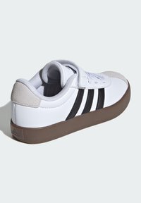 Biały sneaker z czarnymi paskami, beżową łatą na pięcie, zapięciem na rzep i brązową podeszwą, pokazany z tyłu pod kątem na białym tle.