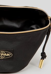 Sac banane en tissu noir avec fermeture éclair dorée et cordon noir avec embouts dorés, avec un petit patch logo Eastpak noir et or.