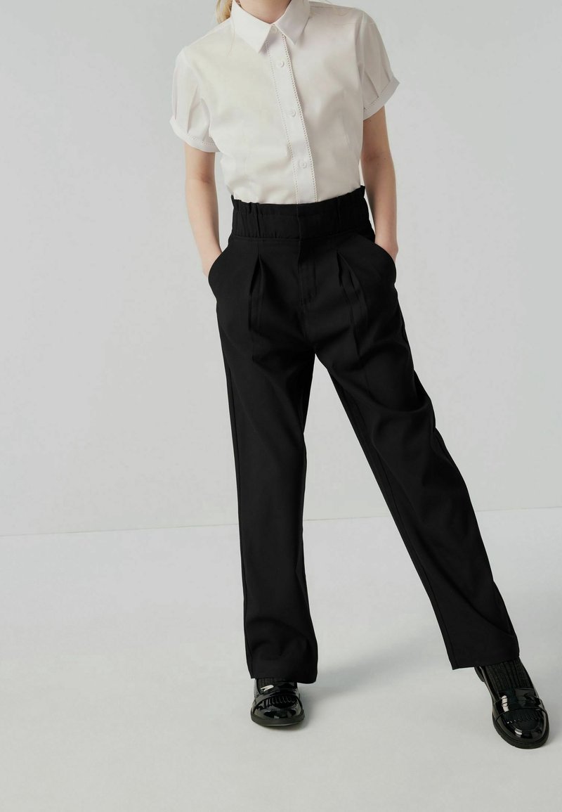 Personne portant une chemise blanche à manches courtes avec col, un pantalon noir plissé taille haute avec les mains dans les poches, et des chaussures noires brillantes.