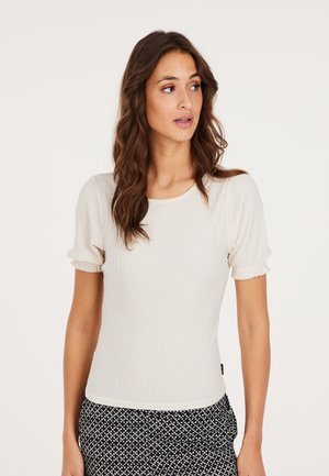 Lys beige ribbet top med korte puffærmer og rund halsudskæring, fremstillet af textured stof; parret med sorte mønstrede bukser.