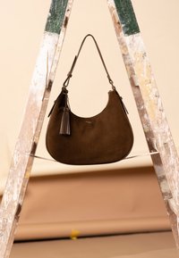 Braune Wildleder-Hobo-Tasche mit geschwungener Form, Quastenakzent und verstellbarem Riemen, präsentiert auf einer bemalten Leiter vor neutralem Hintergrund.