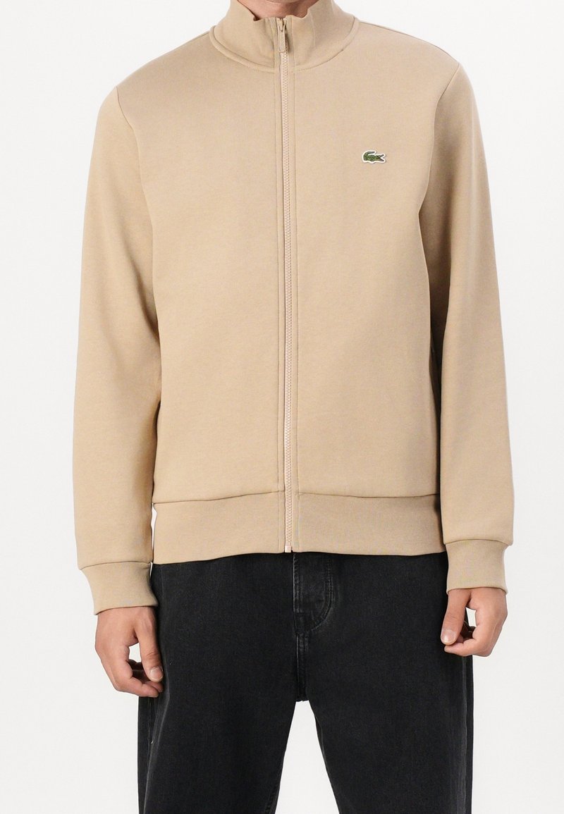 Veste beige à fermeture éclair avec manches longues, petit logo crocodile vert sur la poitrine gauche, portée avec un pantalon noir.