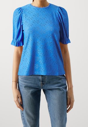 Frau trägt eine leuchtend blaue Bluse aus Lochspitze mit Puffärmeln und gerafften Manschetten, kombiniert mit mittelblauen Jeans.