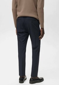 Pantalons taillés bleu marine à coupe droite, avec des poches latérales et des poches arrière à double bouton. Portés avec un pull marron et des chaussures noires.