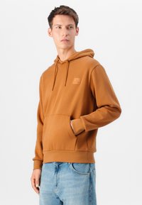 Sweatshirt marron avec une poche kangourou, des cordons ajustables et un logo brodé. Fabriqué en tissu lisse, associé à un jean bleu clair.
