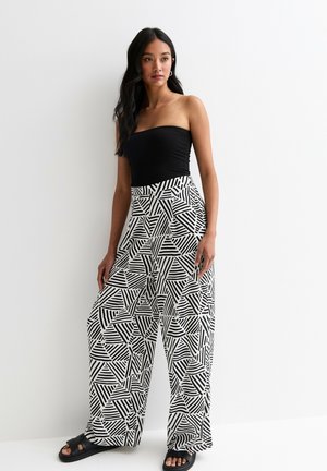 Top negro sin tirantes combinado con pantalones de pierna ancha de cintura alta que presentan un patrón geométrico de rayas en blanco y negro. Sandalias negras de tipo slide.