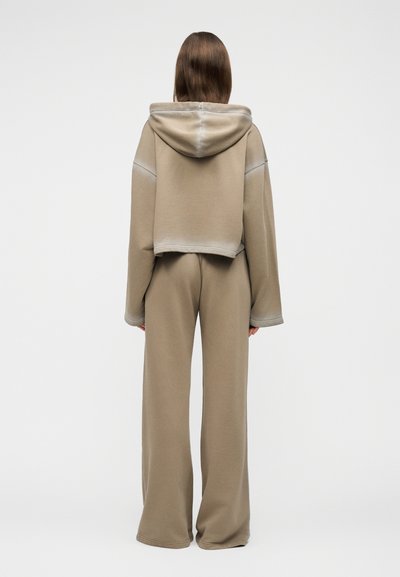 Persona de pie de espaldas, vistiendo una sudadera corta con capucha beige y pantalones de pierna ancha contra un fondo claro y liso.