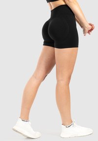 Pantaloni corti neri a vita alta realizzati in tessuto elasticizzato, con un design aderente e una texture liscia, abbinati a scarpe da ginnastica bianche.