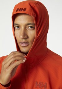 Chaqueta naranja con capucha, que presenta un acabado texturizado, cierre de cremallera y el logo "HH" en un tono más oscuro. Mano ajustando la capucha.