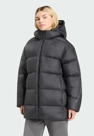 Blonde vrouw in een zwarte donsjas met kap en ritssluiting aan de voorkant, staand met handen dicht bij de zakken tegen een effen achtergrond.